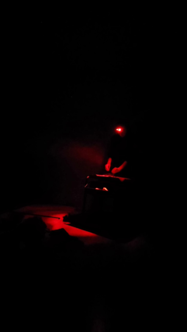 A figure in near-total darkness lit only by a red headlamp, hands working over a low object that catches the light.